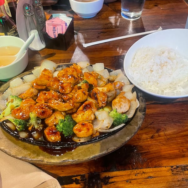 Sizzling teriyaki chicken with broccoli and onions, served with white rice and soup on a wooden table.