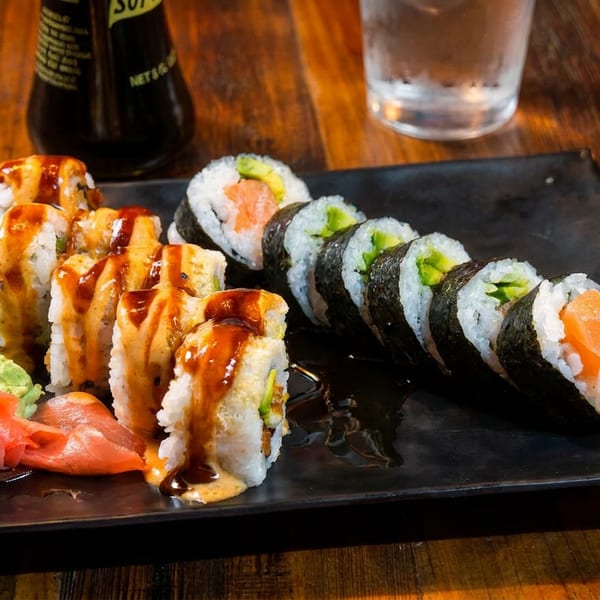 A black plate of sushi rolls with salmon, avocado, wasabi, and pickled ginger on a wooden table.