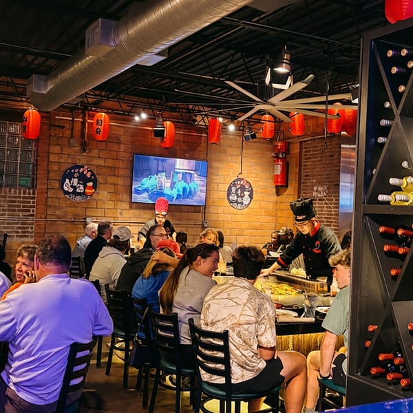 Diners seated around a hibachi grill, watching a chef prepare food in a lively restaurant.