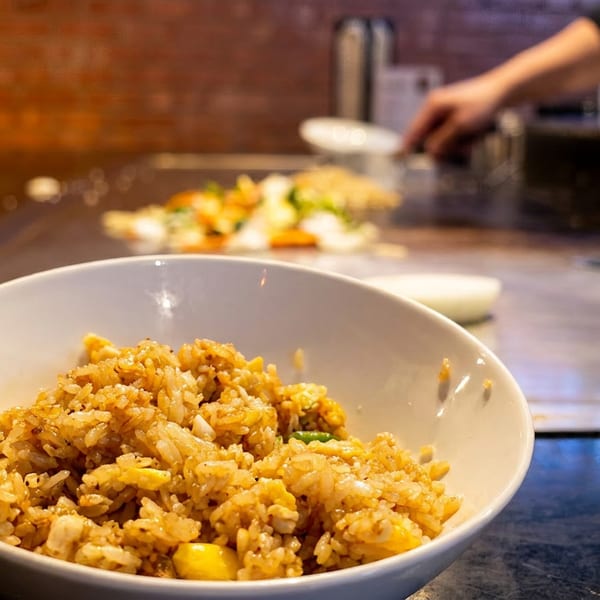 A bowl of fried rice in the foreground with a chef cooking on a hibachi grill in the background.